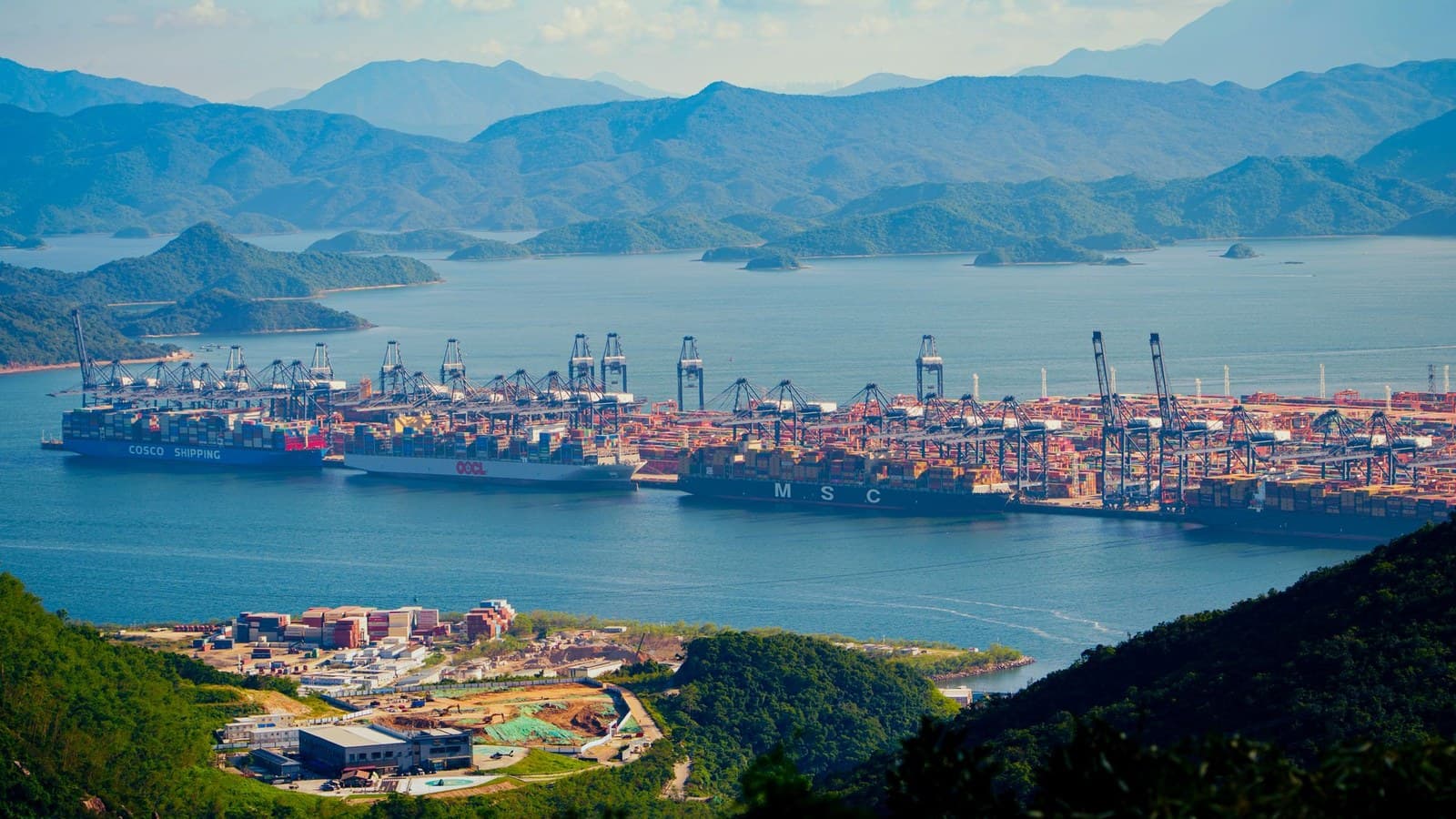 从山坡上俯看一座集装箱码头：岸边是岸桥和成排堆叠的集装箱，船只靠泊，背后是青山。