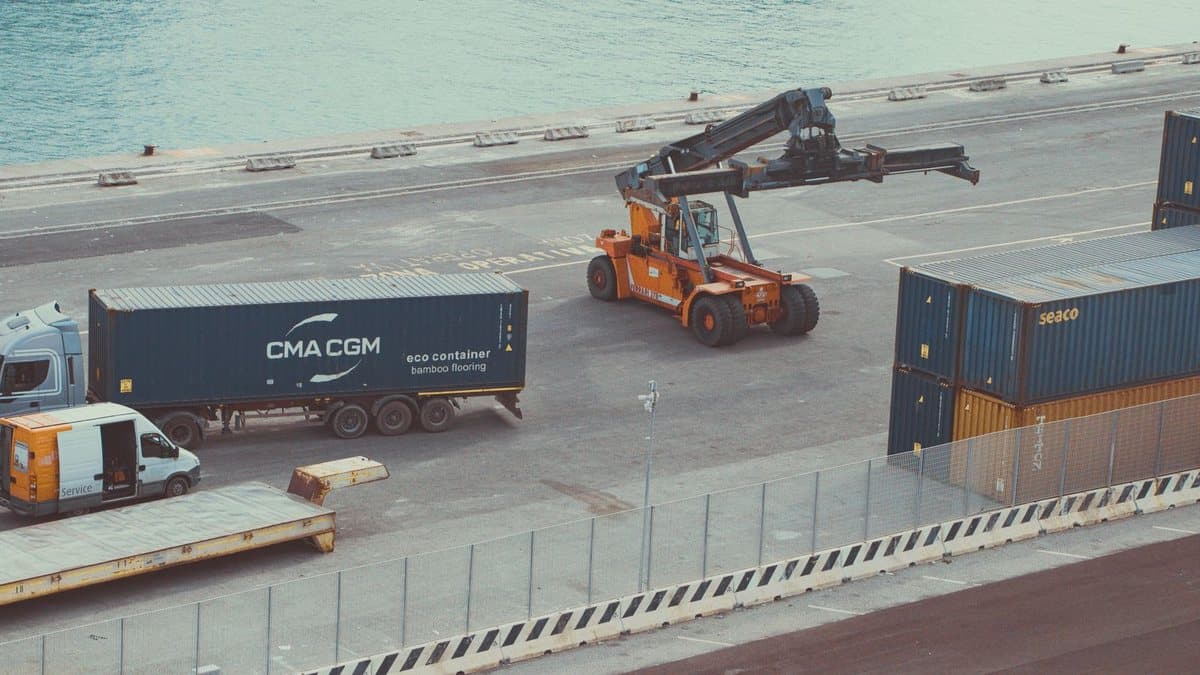 A port terminal shows a truck with a container, a container handling machine, and stacked containers near the water.