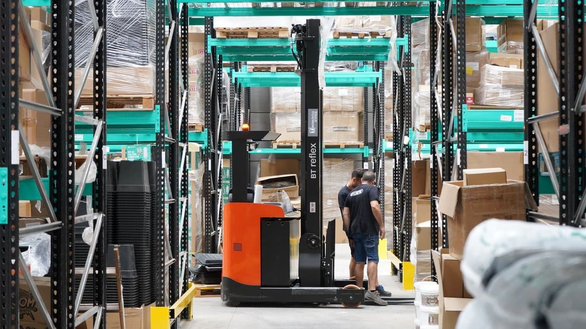 Two workers stand by an orange reach truck in a vast warehouse filled with stacked boxes on tall metal shelves.