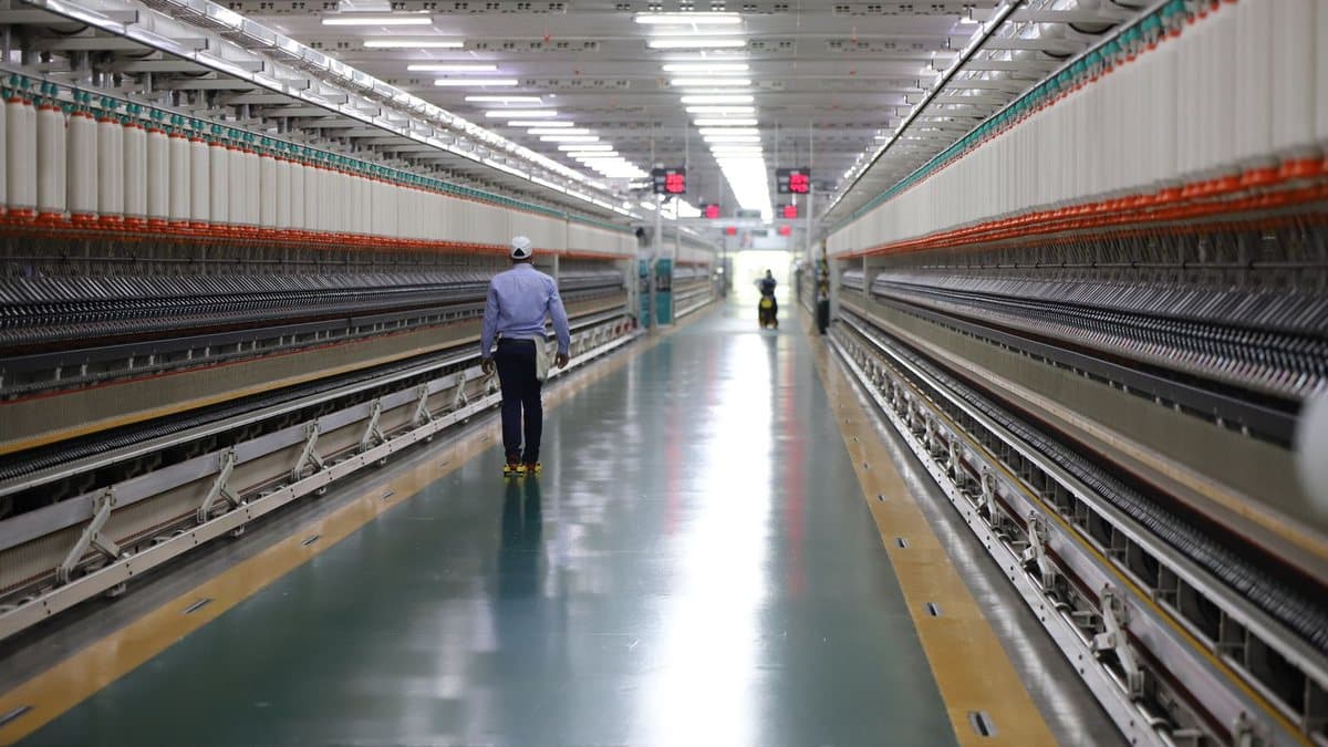 A worker walks down a long, brightly lit factory aisle between rows of textile machinery holding countless white spools of yarn.