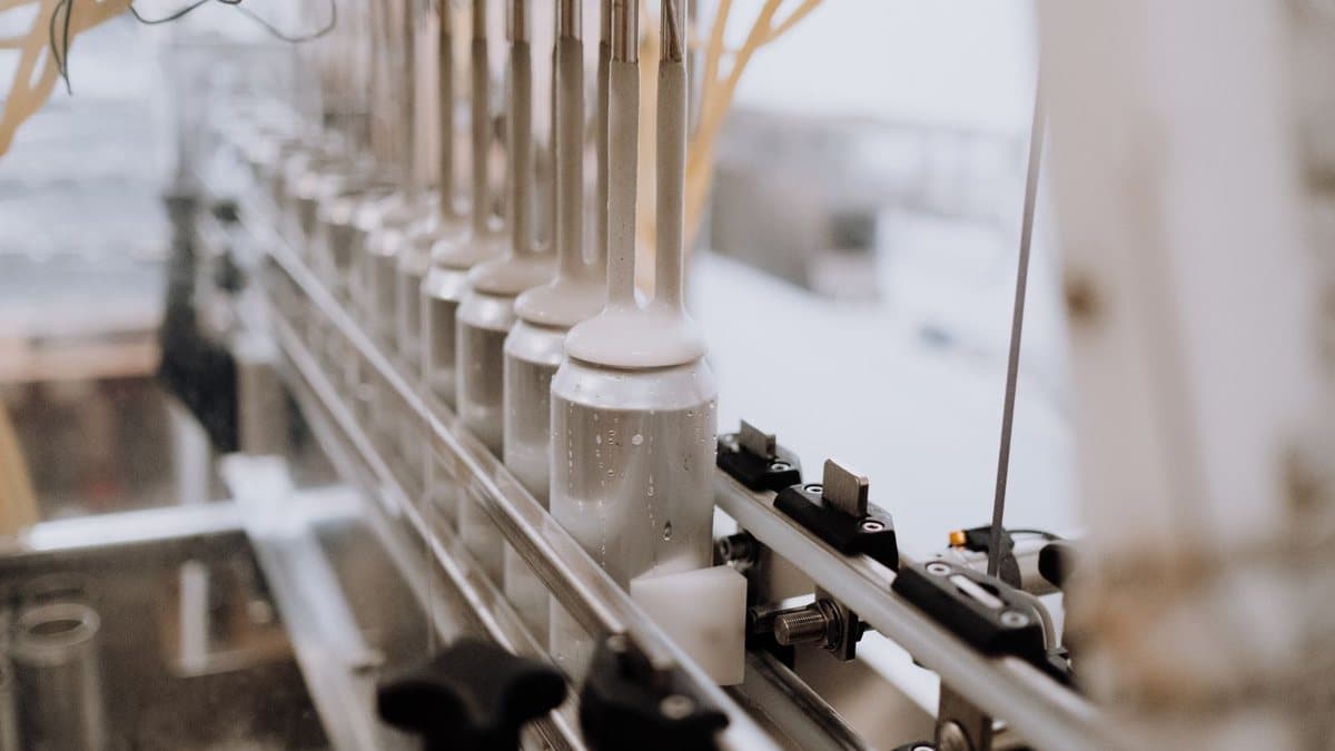 Rows of wet aluminum cans are being filled by multiple nozzles on a conveyor belt in a bright factory.