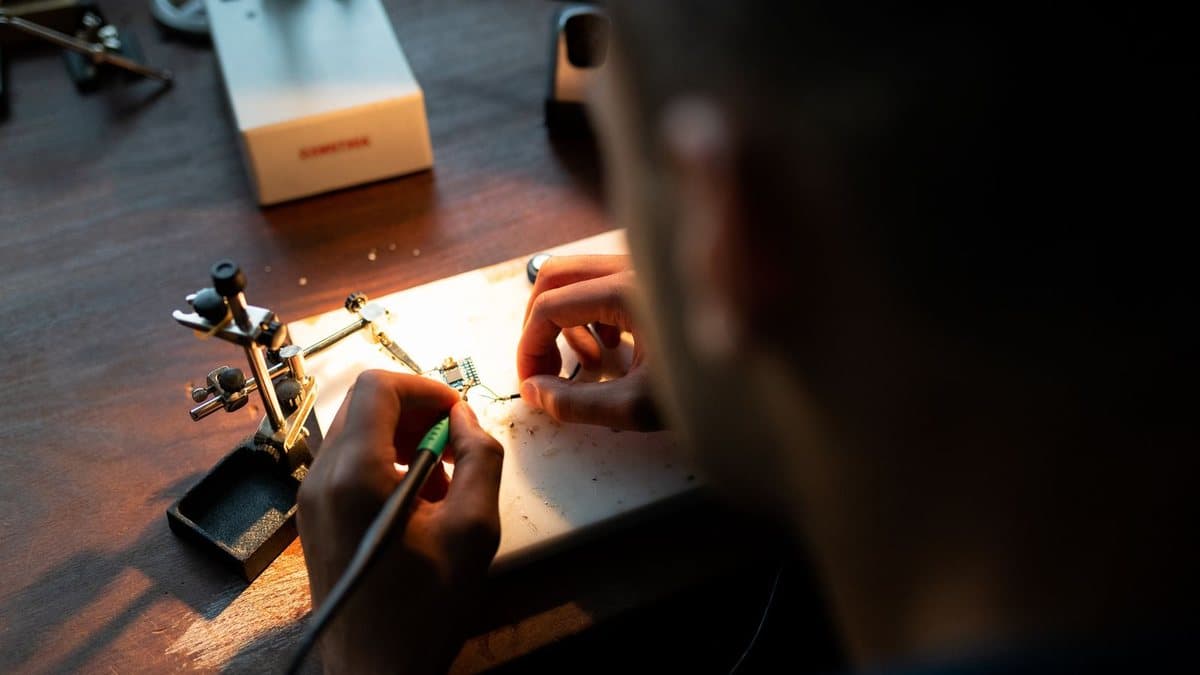 A person's hands are soldering a small circuit board held by a helping hands tool on a wooden desk.
