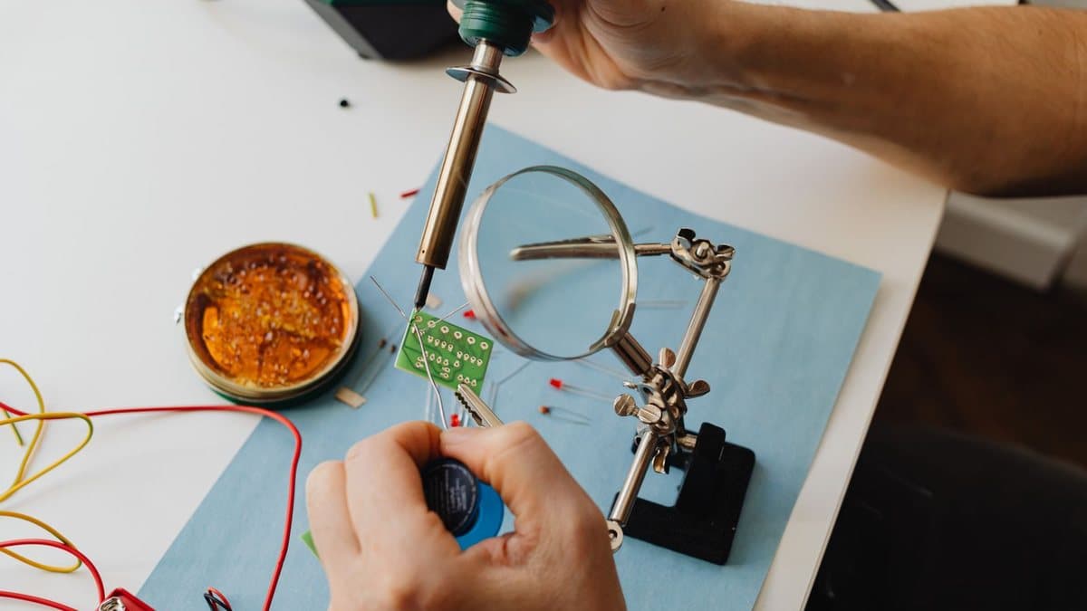 Hands solder electronic components onto a green circuit board on a workbench, using a soldering iron and magnifying glass.