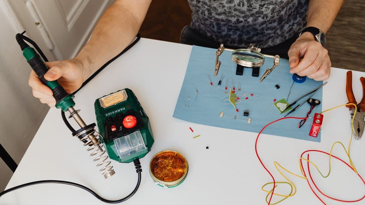 A person solders electronic components on a white table, surrounded by tools and various circuit parts.
