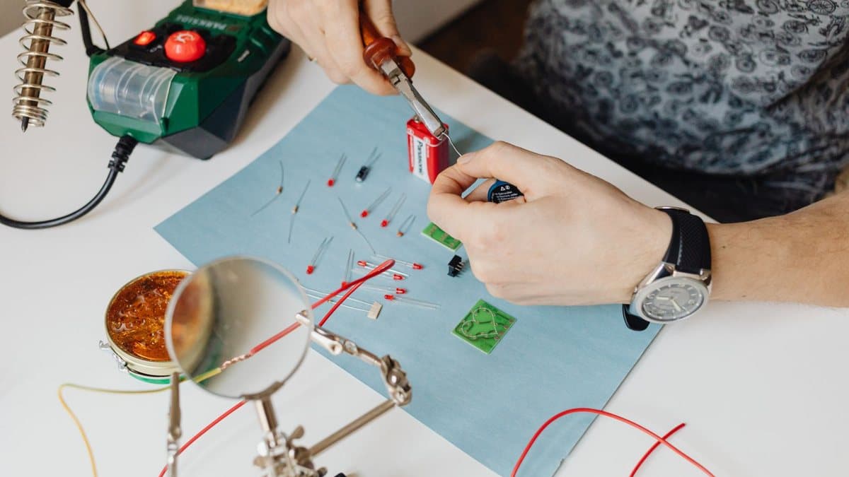 A person's hands assemble small electronic components like LEDs and circuit boards on a table with a soldering station and tools.