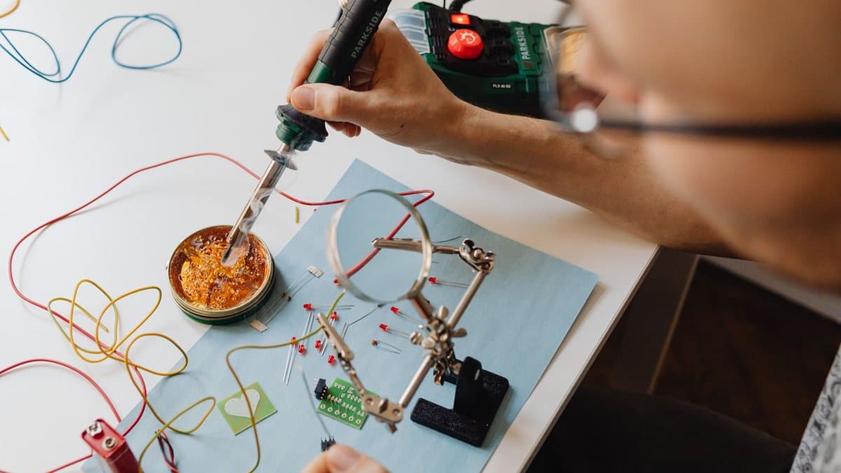 A person wearing glasses uses a soldering iron to melt solder, surrounded by electronic components and wires.