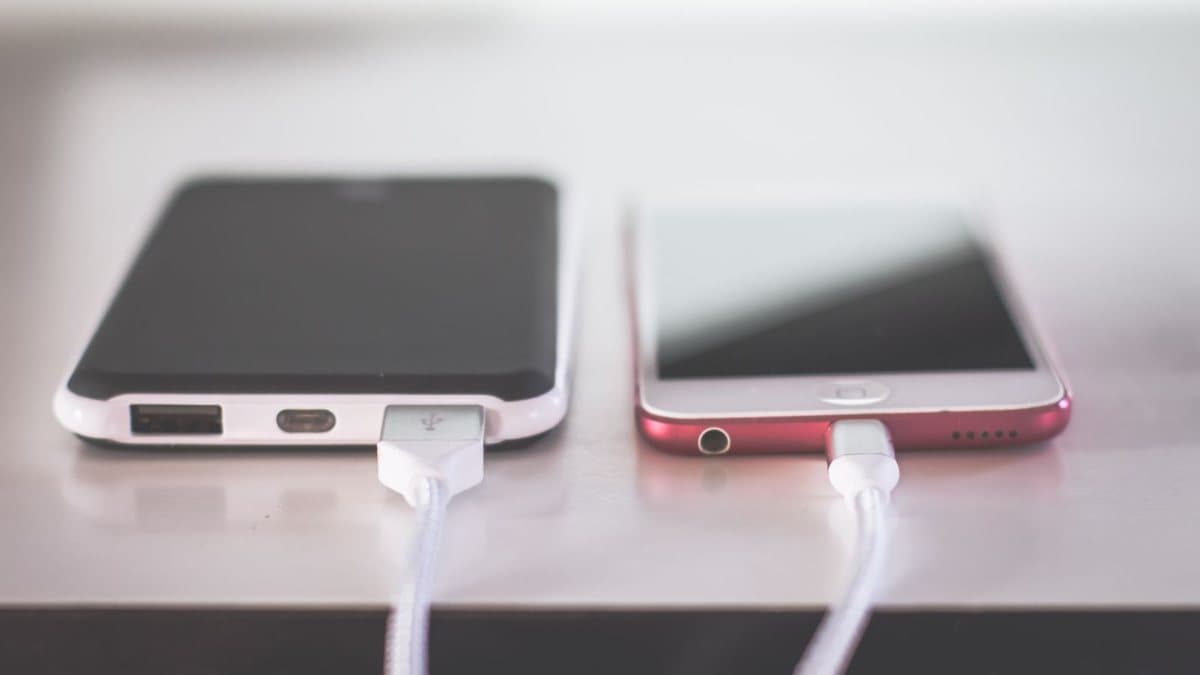 Two mobile phones are connected by a white cable, with the left phone charging the right phone on a light-colored surface.