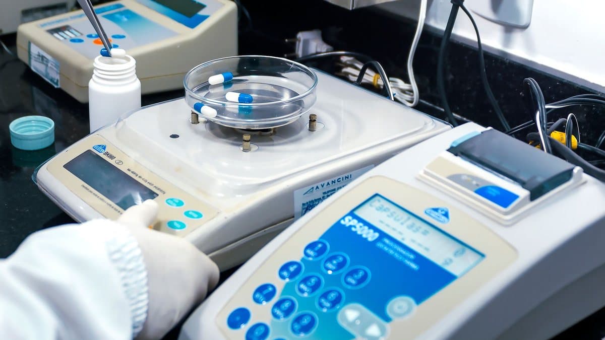 Two gloved hands operate lab equipment, one placing a capsule into a bottle with tweezers, the other adjusting a digital scale.