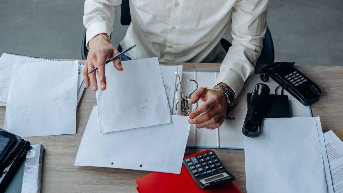 A person in a white shirt sorts papers into a binder on a wooden desk, with a phone and calculator.