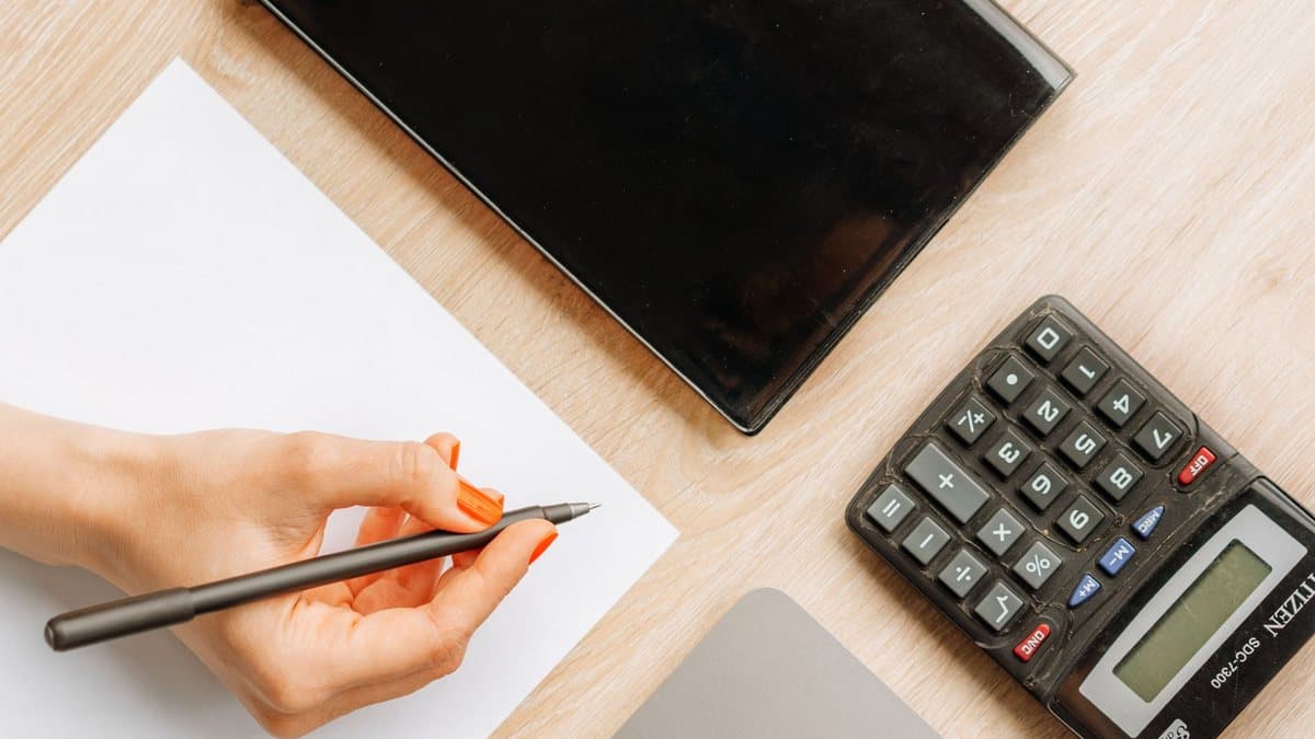 A hand with orange nail polish holds a pen over a white paper, next to a black tablet and a calculator on a wooden desk.