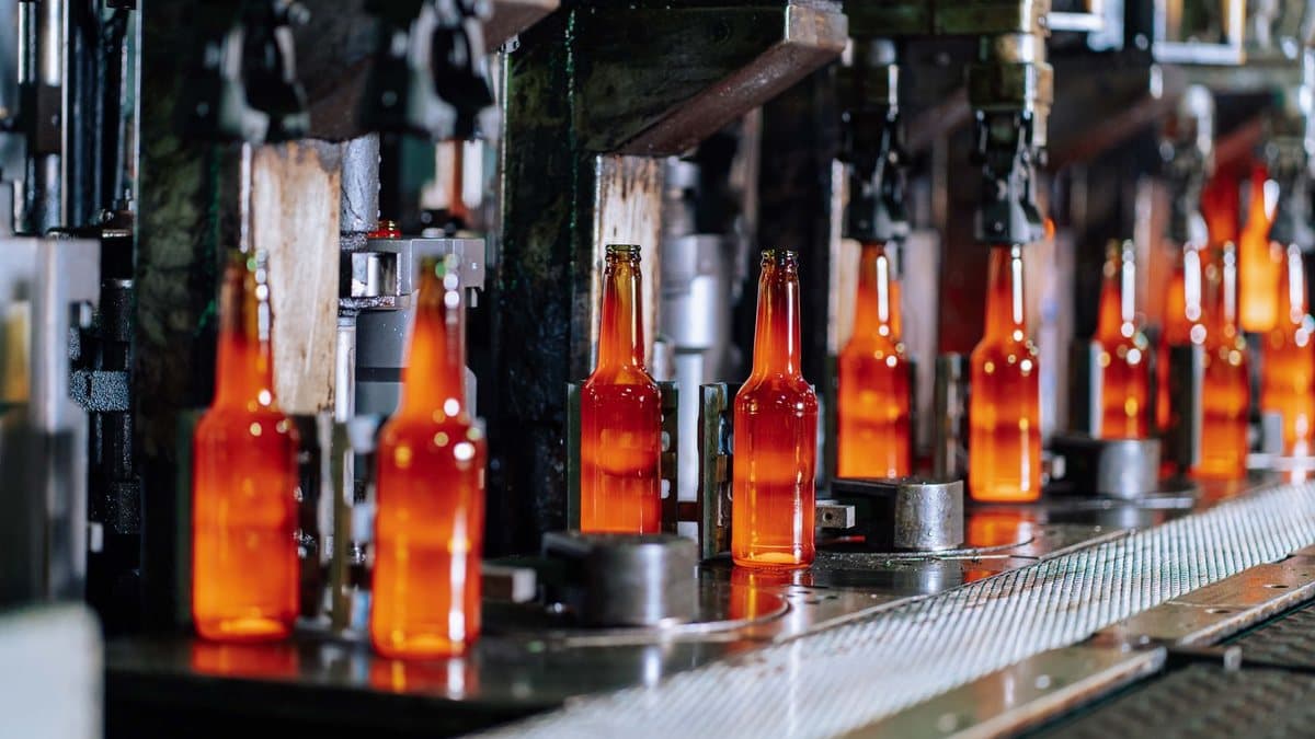 Rows of hot, glowing orange glass bottles move along a conveyor belt in an industrial facility.