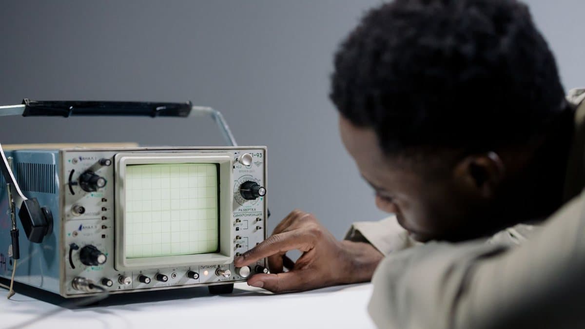 A person leans over a vintage oscilloscope, intently adjusting its controls with one hand.