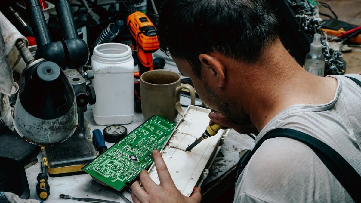 A person solders an electronic circuit board on a cluttered workbench amidst various tools and components.