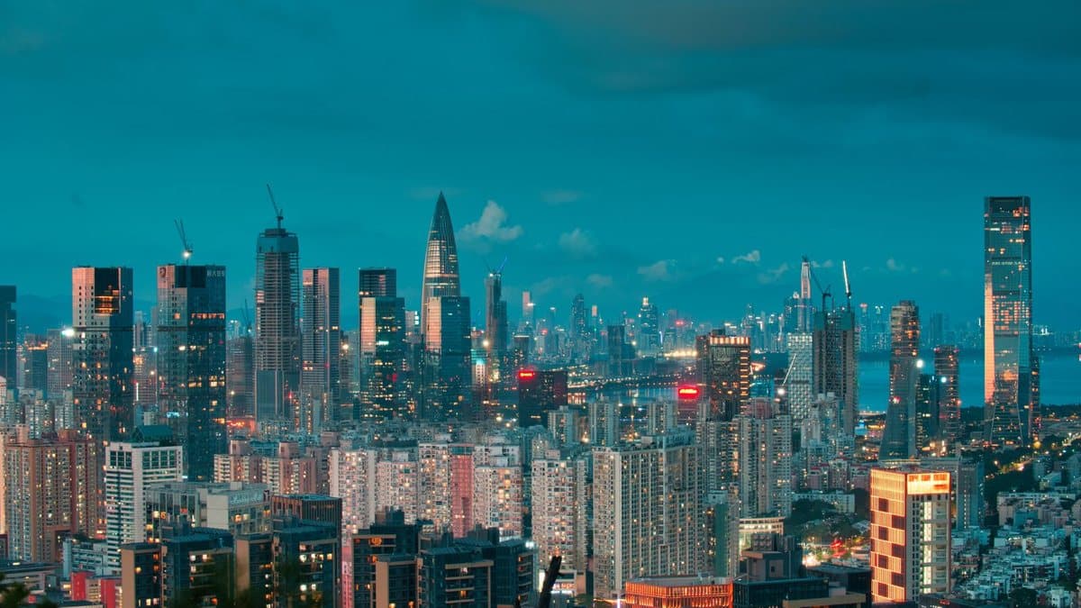 黄昏时分,广阔的现代都市灯火璀璨,高楼林立,远处的蓝色水域映衬在青色天幕下。