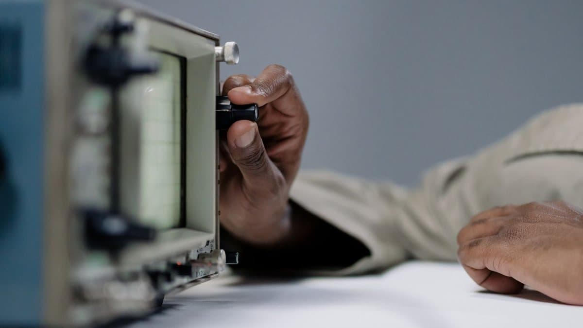 A dark-skinned hand adjusts a knob on a piece of electronic test equipment with a grid display, sitting on a white table.