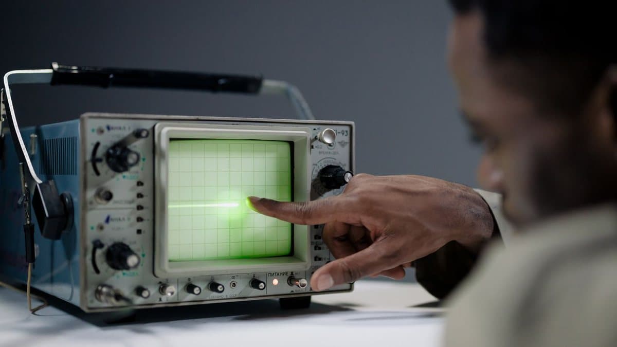 A person's hand points to a green glowing line on the screen of an old oscilloscope, set in an indoor environment.