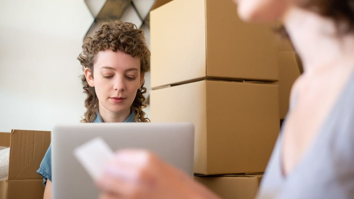 A woman with curly hair uses a laptop while another woman holds a card, with many cardboard boxes stacked nearby.