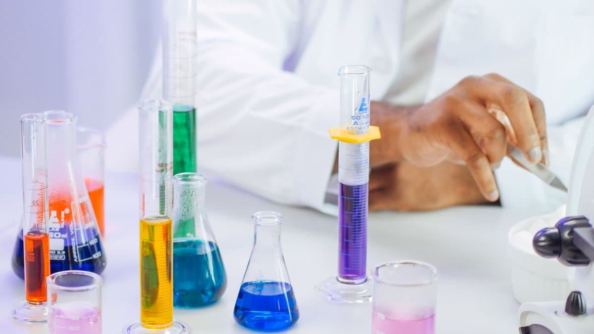 A person in a white lab coat uses a tool near a microscope, surrounded by various glass beakers and flasks filled with colorful chemical liquids.