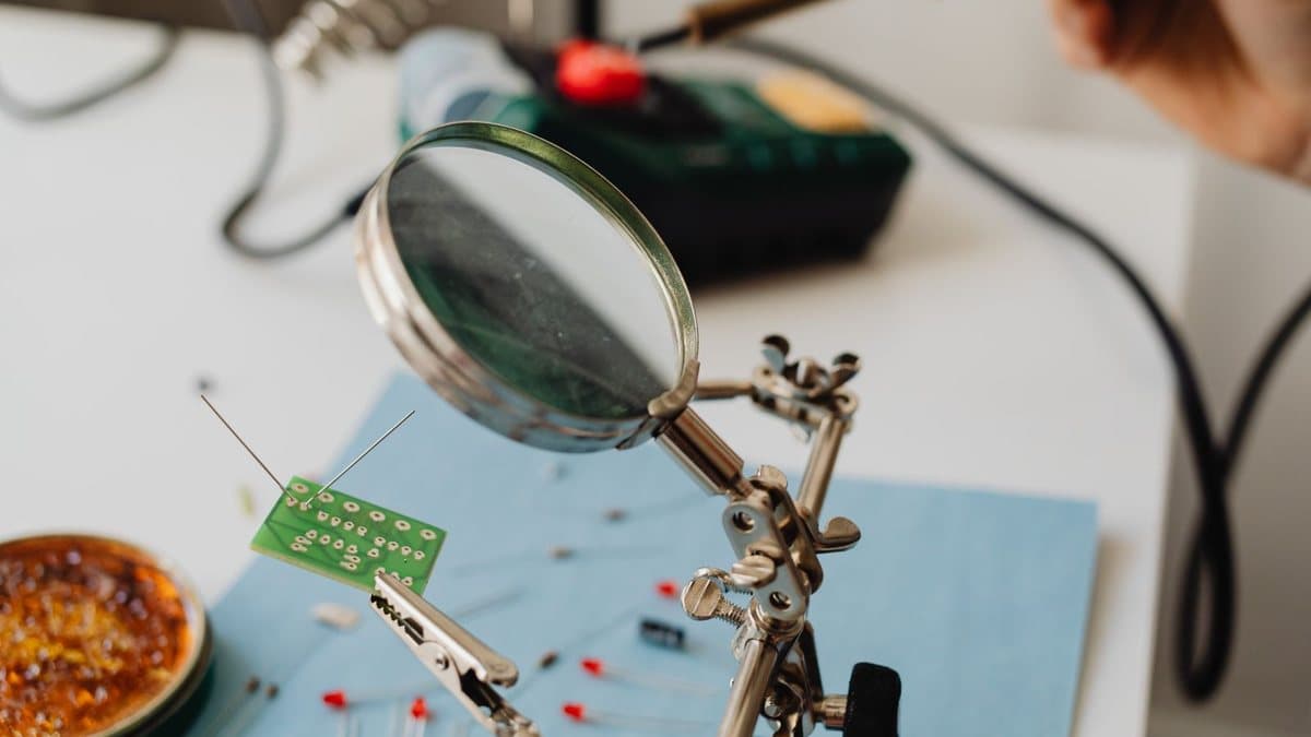 A person solders electronic components onto a green circuit board, using a helping hands tool with a magnifying glass.