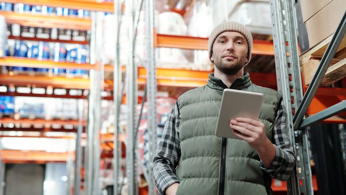 A man in a beanie and vest stands in a warehouse aisle, holding a tablet and looking forward, with blurred storage shelves in the background.
