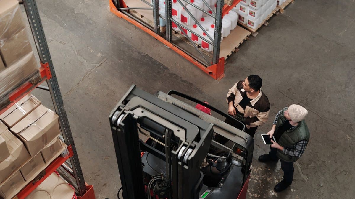 Two workers stand by a forklift, observing tall warehouse racks filled with boxes and white containers.