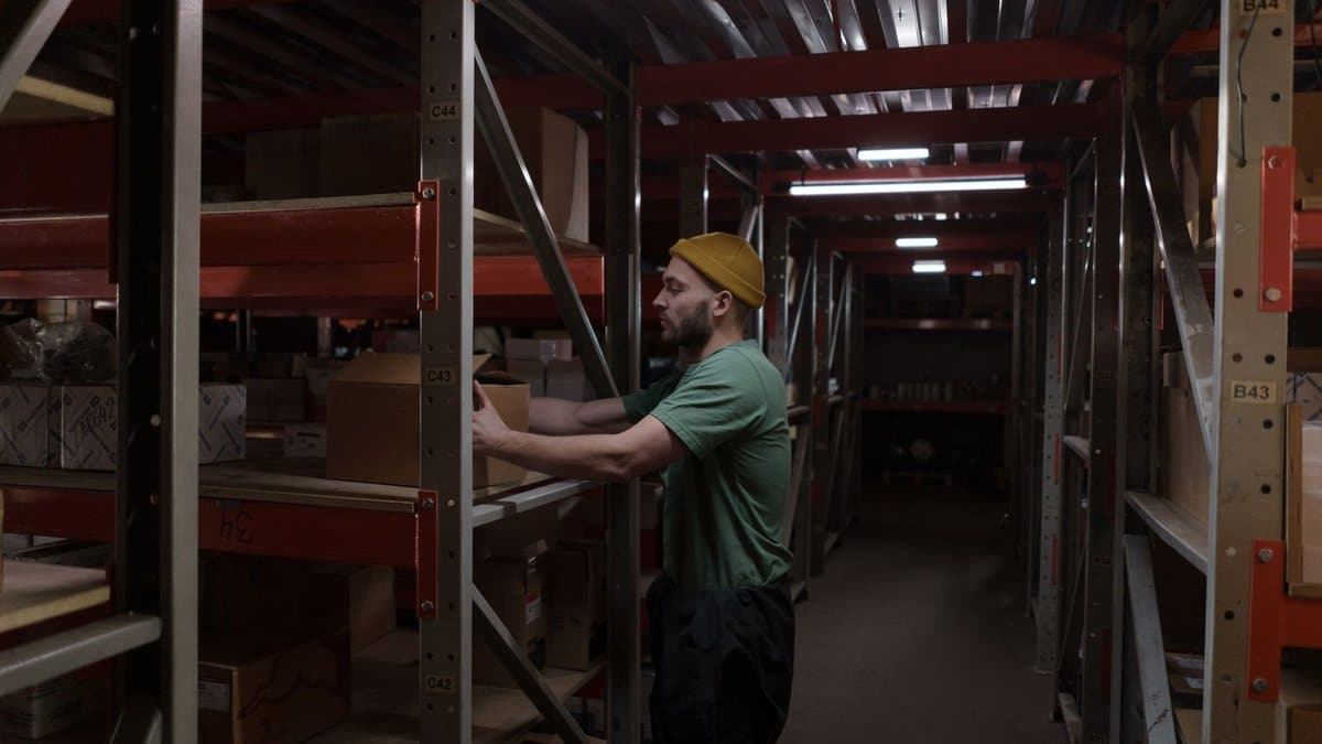 A man in a yellow beanie and green shirt places a cardboard box on a shelf in a dimly lit warehouse aisle filled with inventory.