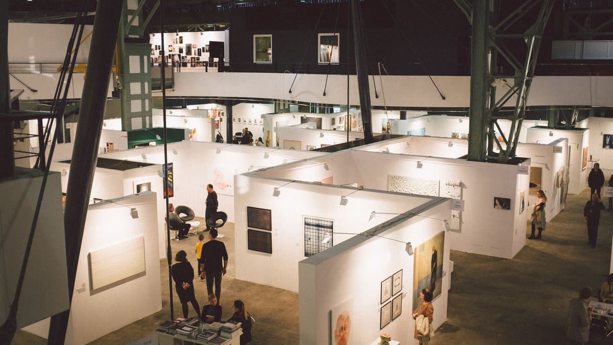 An aerial view of an indoor art exhibition with multiple white booths displaying artworks and numerous visitors walking through the maze-like space.
