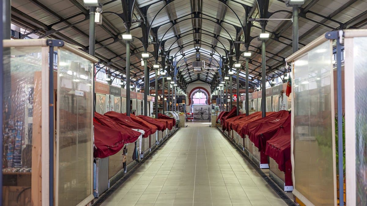 An empty, well-lit indoor market hall with stalls covered in red cloths lining a tiled aisle, leading to an arched window.