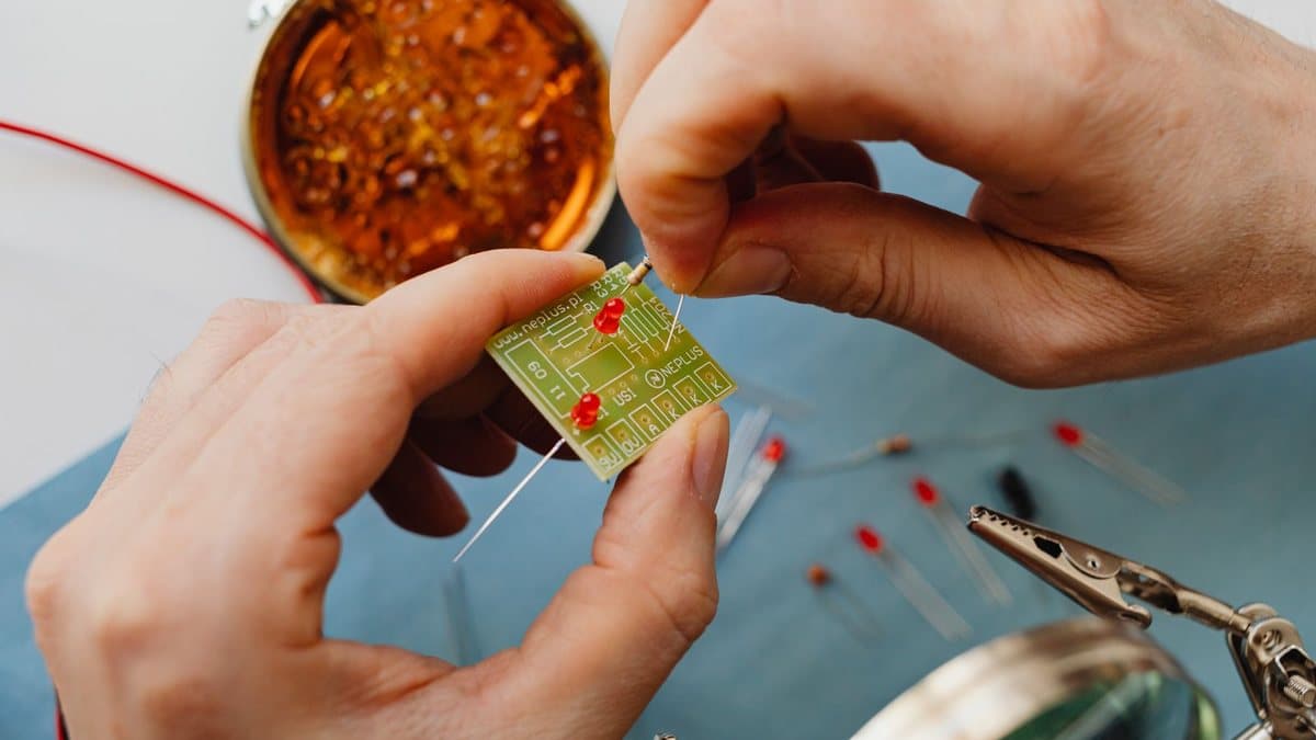 一双手正在将一个电阻安装到一块小型绿色电路板上,桌上还有其他电子元件和工具。