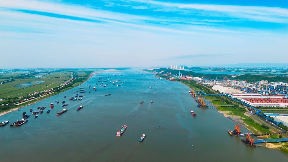 An industrial complex and agricultural fields flank a busy waterway filled with numerous cargo vessels under a partly cloudy sky.