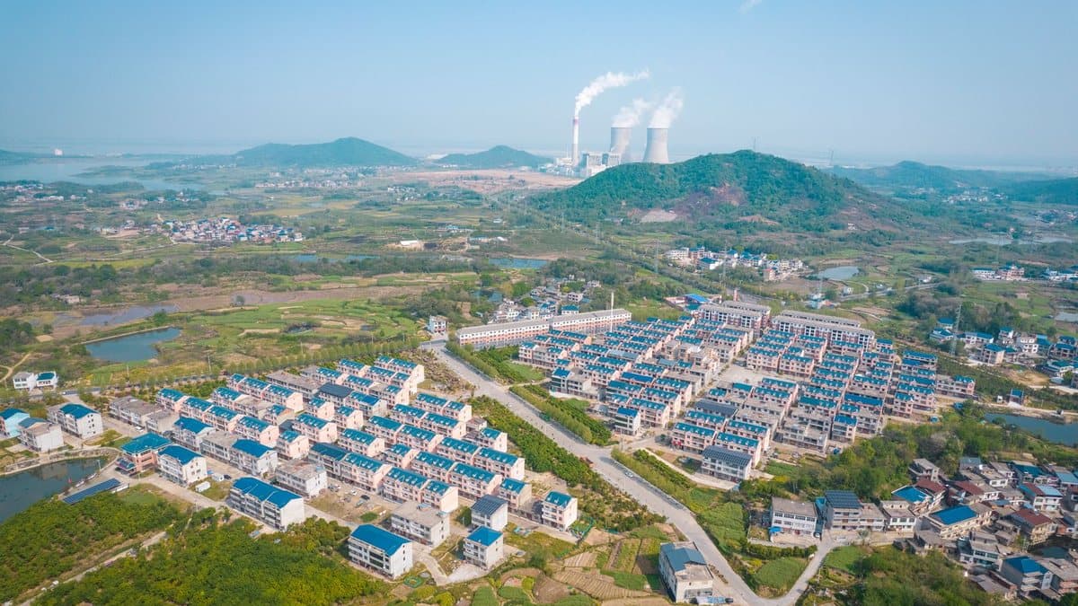 Rows of uniform houses with blue roofs fill a green landscape, with a power generation facility emitting smoke and steam in the background.