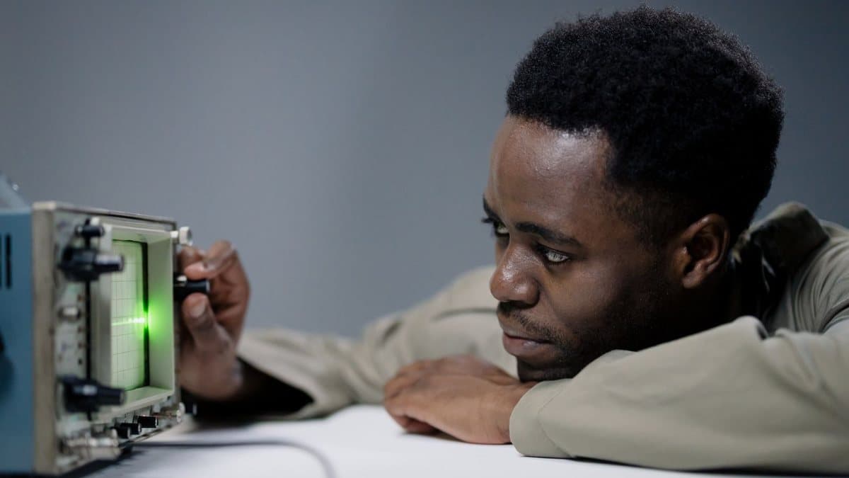 A man intently adjusts a knob on an oscilloscope showing a green waveform, resting his forearms on a white table.