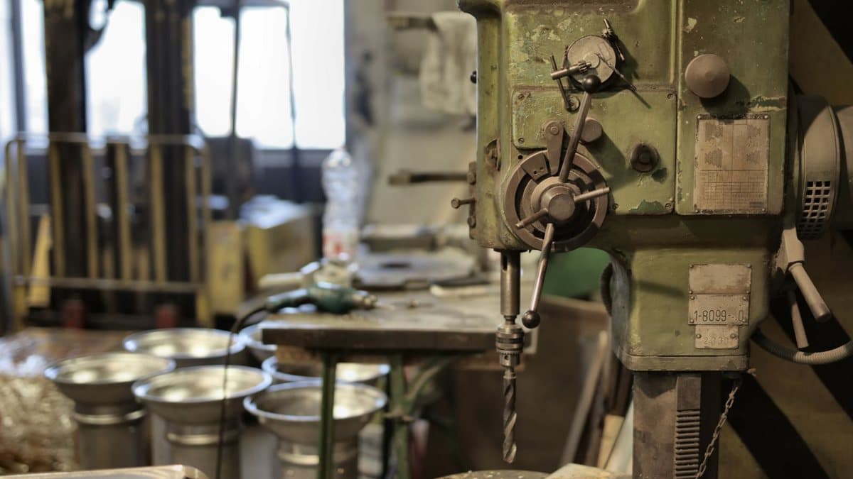 An old, green industrial drilling machine with a drill bit, surrounded by tools and metal containers in a workshop.