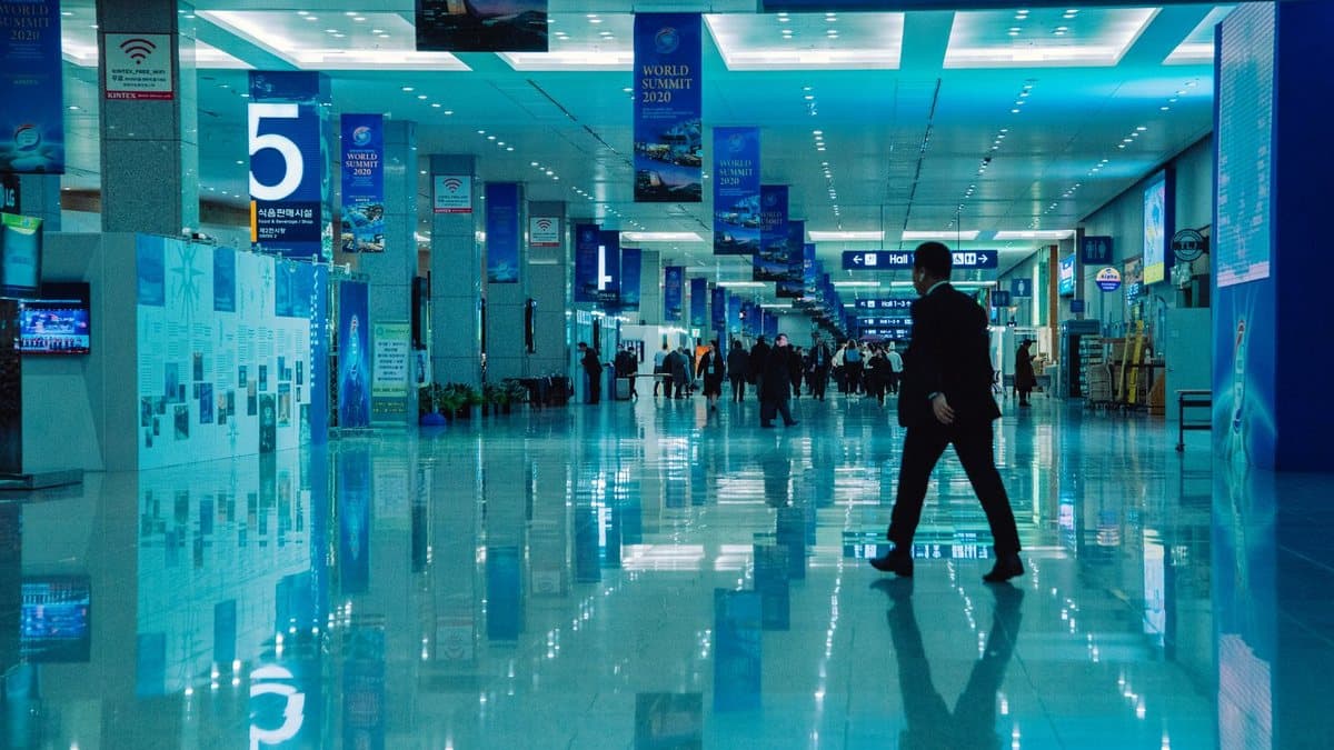 A wide, blue-lit hall features reflective floors and informational banners, with people walking and a man in a dark suit in the foreground.