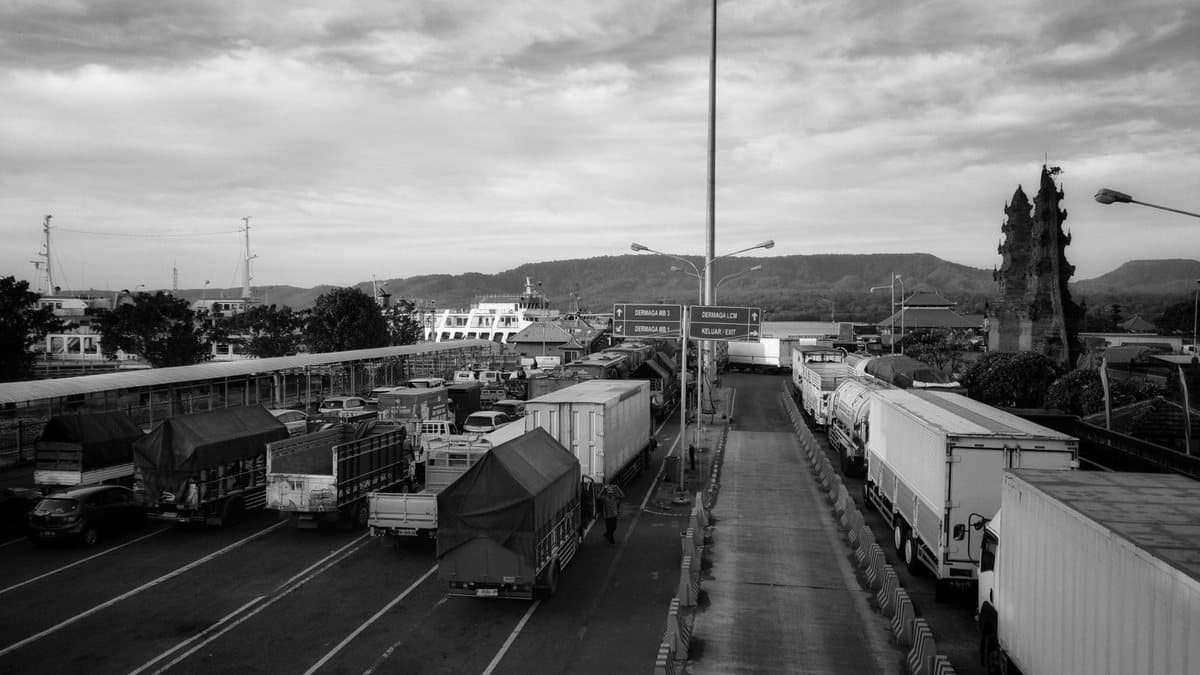A busy black and white port scene shows trucks and cars queuing for ferries, with a distinctive tiered temple gate nearby.