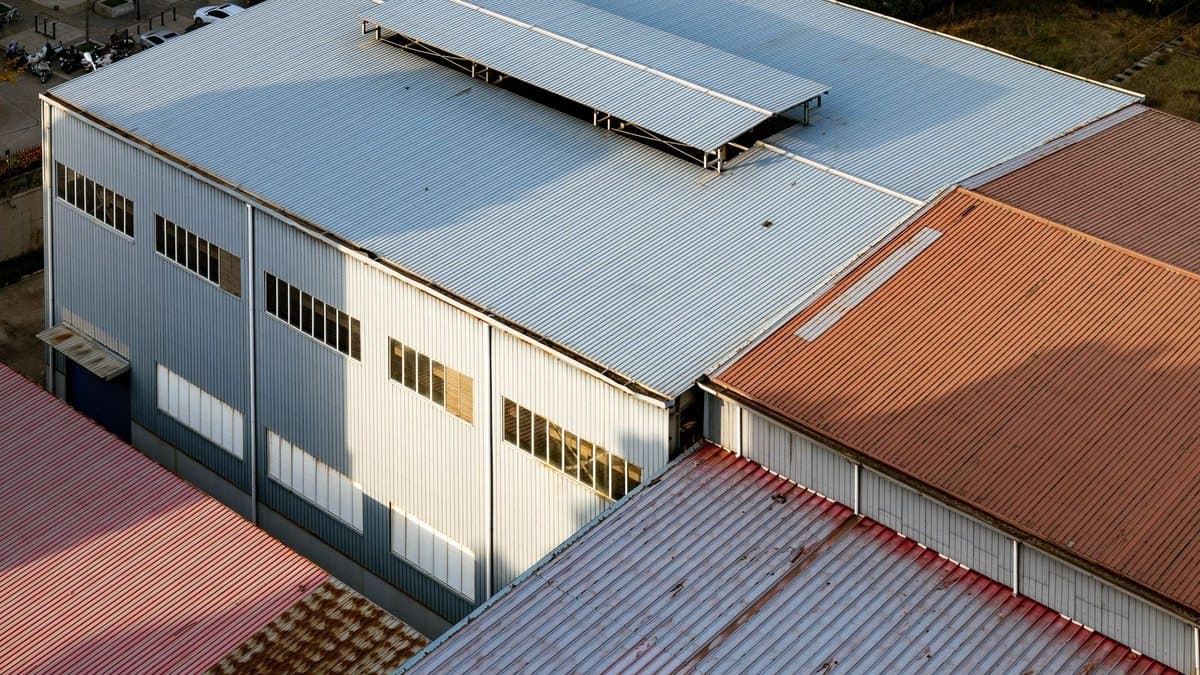 An aerial view of multiple industrial buildings with corrugated metal roofs and walls, casting shadows under bright sunlight.
