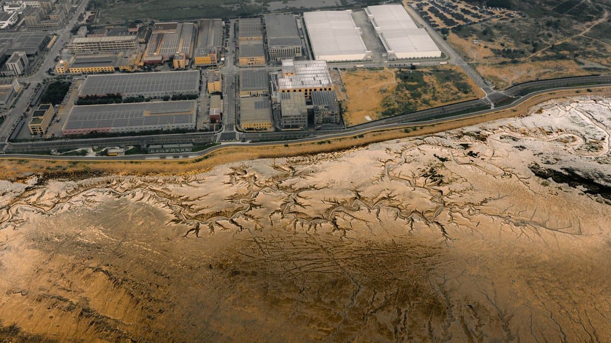 An aerial view shows an industrial complex with many buildings and roads next to expansive, dry mudflats with intricate natural patterns.