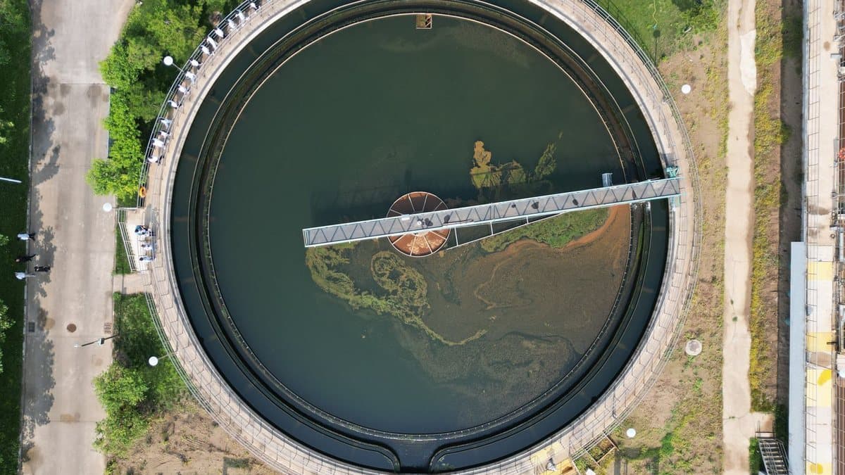 An aerial view shows a large circular water treatment tank with a central walkway, surrounded by paved and grassy areas, where several people are observing.