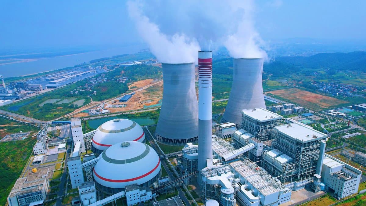 An aerial view shows a vast power plant with tall cooling towers, a chimney, and large storage domes, set by a wide river and green landscape.