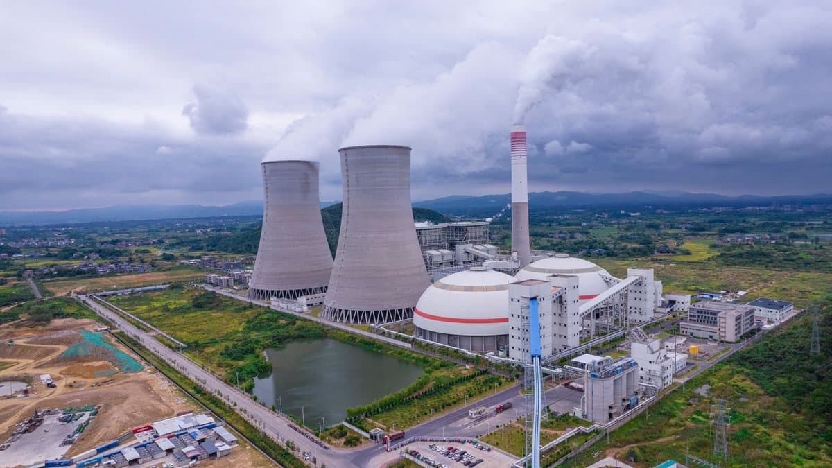 Two large cooling towers and a tall smokestack at an industrial complex emit vapor into a cloudy sky, surrounded by green landscape.
