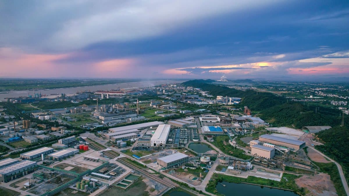 An extensive industrial complex with numerous buildings and a winding river, bordered by hills, under a vibrant sunset sky with clouds.