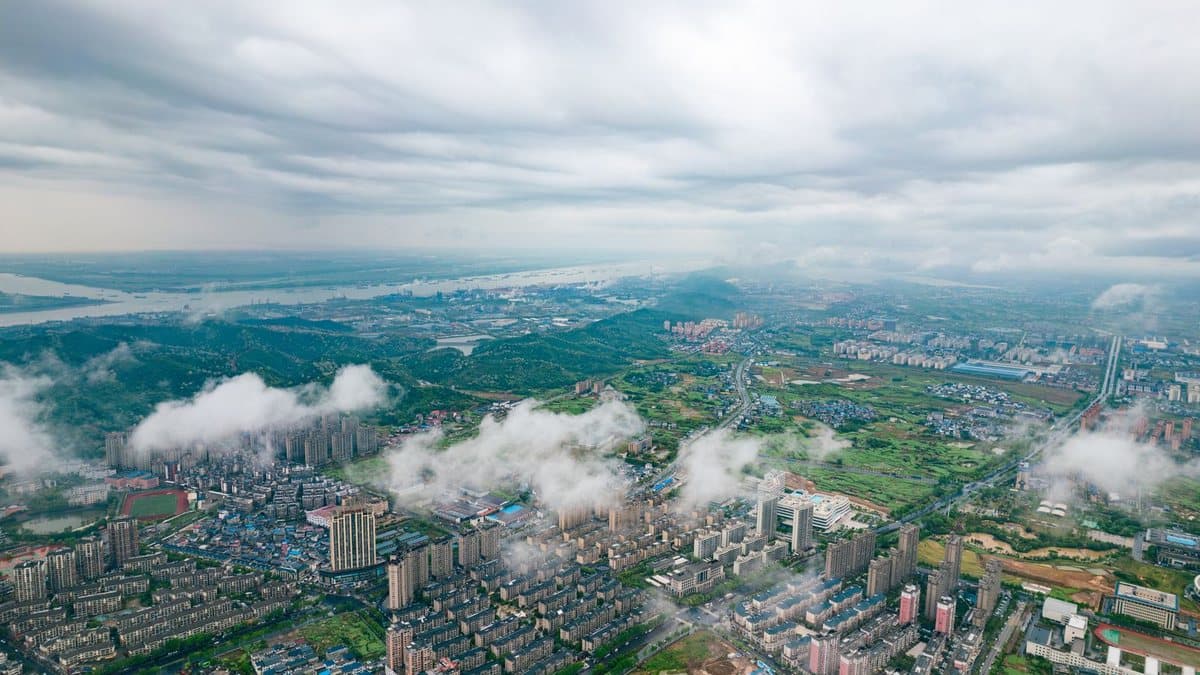 An expansive aerial view reveals a city with numerous buildings, green areas, a wide river, and misty hills under a cloudy sky.