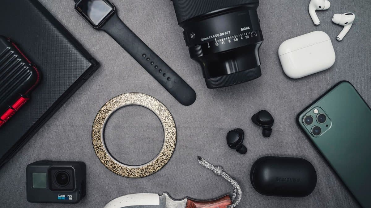 Various tech gadgets including a smartphone, cameras, wireless earbuds, and a smart watch are arranged on a gray fabric surface, alongside a decorative metal ring and a knife.