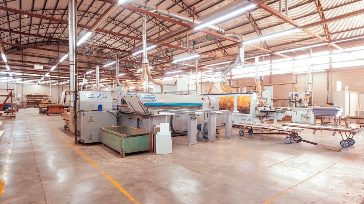 A well-lit, spacious woodworking factory floor shows large panel processing machines, dust collection systems, and organized wood materials.