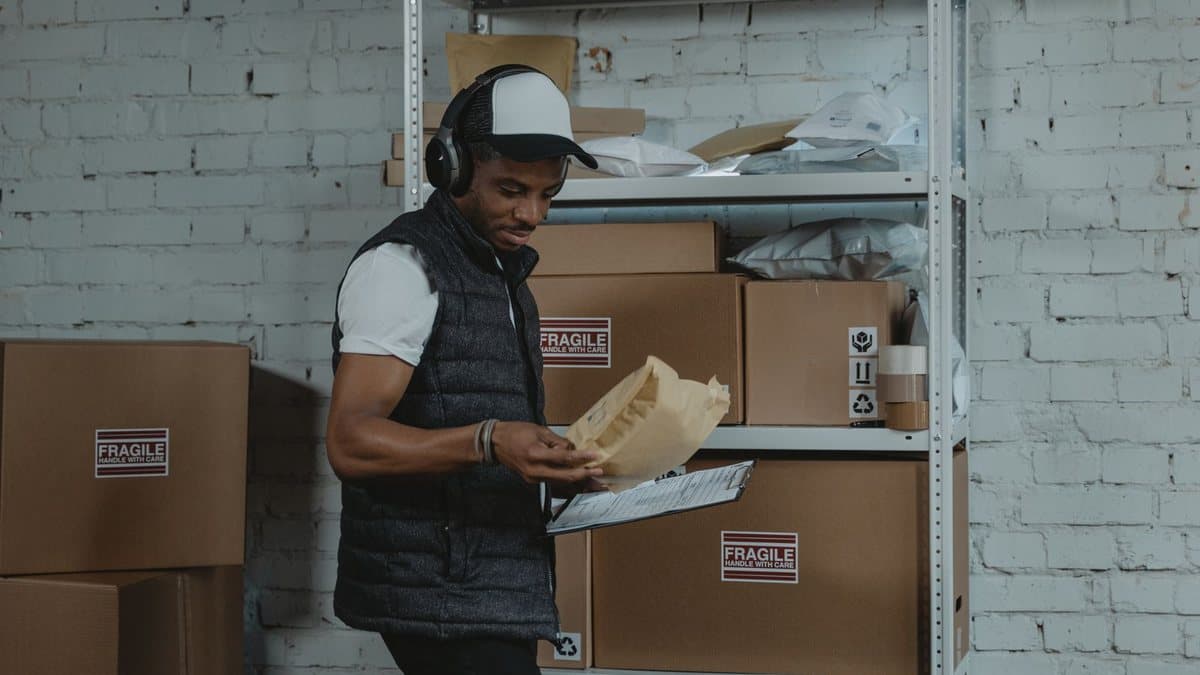A man wearing headphones and a vest checks a package and a clipboard in a storage area with shelves and boxes.