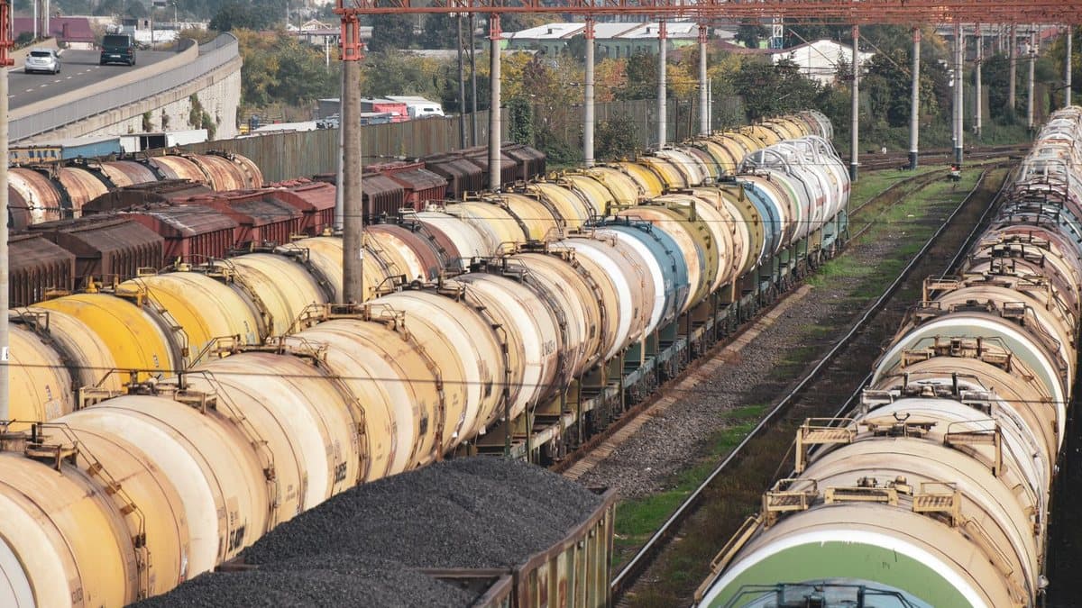 Rows of tank cars and freight wagons, some filled with coal, are parked on multiple railway tracks next to an elevated highway.