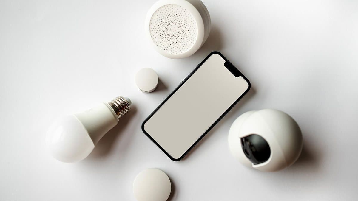 Smart home devices including a smartphone, light bulb, security camera, speaker, and two small sensors on a white surface.