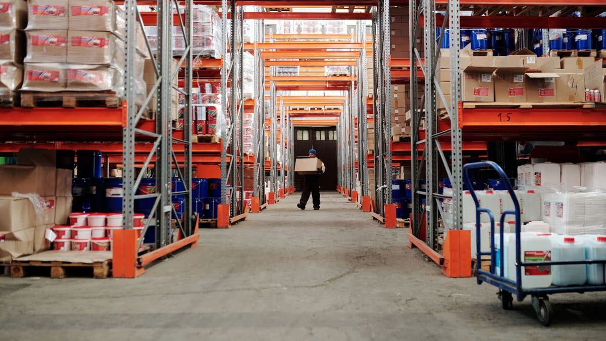 A person walks down a long aisle in a well-stocked warehouse, carrying a cardboard box past rows of loaded shelves.