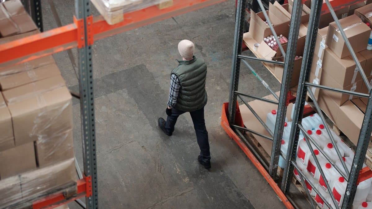 An overhead view shows a person wearing a vest and beanie walking on a concrete floor in a warehouse surrounded by tall racks filled with boxes and bottles.