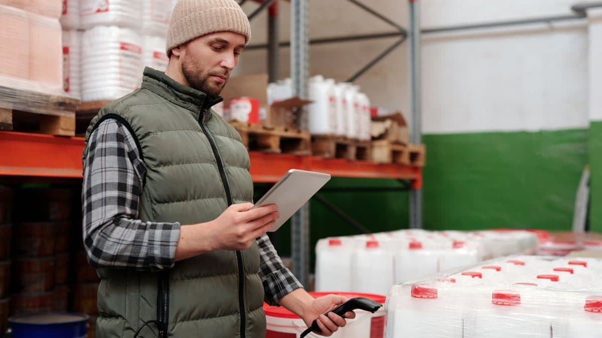 A man in a beanie and padded vest uses a barcode scanner on stacked containers while viewing a tablet in a warehouse.